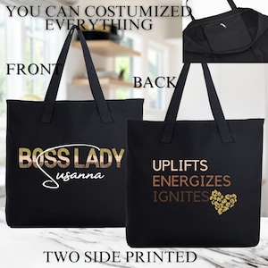 May include: Black canvas tote bag with two-sided printing. The front side features the text "Boss Lady" with a gold glitter design and the name "Susannah" written underneath. The back side features the text "Uplifts Energizes Ignites" with a gold glitter heart design.