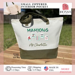 May include: A white canvas tote bag with a green bottom. The bag has a green strap and a small zippered pocket. The bag has the text "MAHJONG" and "Mr. Charlotte" printed on it. There are also images of mahjong tiles printed on the bag.