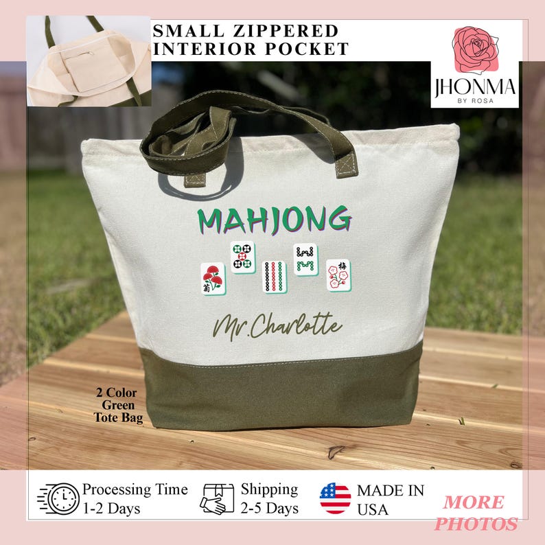 May include: A white canvas tote bag with a green bottom. The bag has a green strap and a small zippered pocket. The bag has the text "MAHJONG" and "Mr. Charlotte" printed on it. There are also images of mahjong tiles printed on the bag.