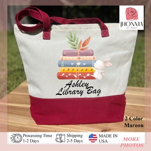 Op de afbeelding: Een witte canvas tote bag met een maronkleurige rand. De tas heeft een stapel boeken met een wit konijn erop. De tekst "Ashley Library Bag" staat op de tas gedrukt.