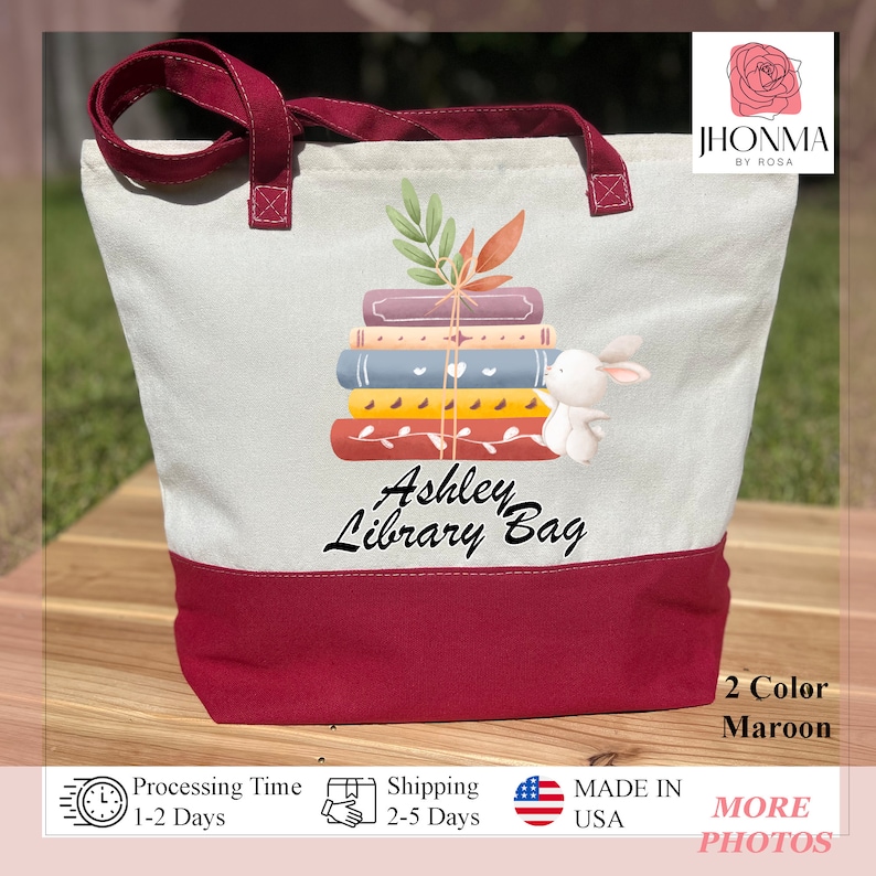 Op de afbeelding: Een witte canvas tote bag met een maronkleurige rand. De tas heeft een stapel boeken met een wit konijn erop. De tekst "Ashley Library Bag" staat op de tas gedrukt.