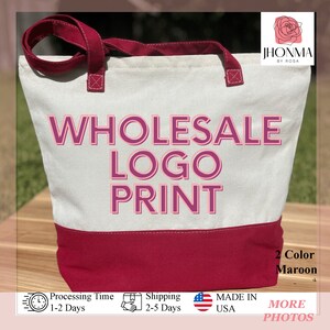 Pu&ograve; includere: Una borsa tote in tela bianca con bordi e manici marroni. La borsa ha il testo "WHOLESALE LOGO PRINT" in lettere rosa. La borsa &egrave; appoggiata su una superficie di legno.