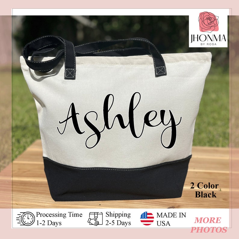 May include: A white canvas tote bag with black handles and a black bottom. The bag has the name "Ashley" written in black cursive on the front. The bag is made in the USA.