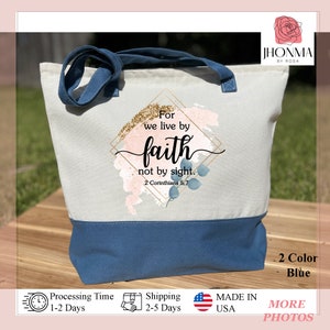 以下が含まれることがあります： 「For we live by faith not by sight. 2 Corinthians 5:7」という文字が印刷された青と白のトートバッグ。バッグは水彩画のデザインで、金色のグリッターのアクセントが施されています。