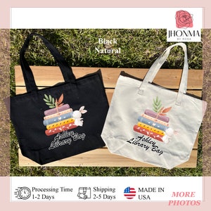 Op de afbeelding: Twee tote bags, een zwarte en een natuurlijke, met de tekst "Ashley's Library Bag" en een stapel boeken met een konijn erop.