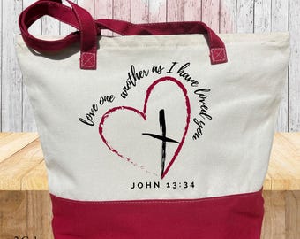 Puede incluir: Una bolsa de lona blanca con asas y fondo marrones. La bolsa tiene un corazón rojo con una cruz negra en su interior y el texto "Love one another as I have loved you" y "John 13:34".