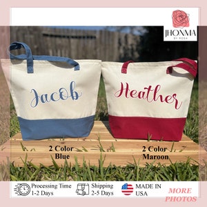 May include: Two personalized canvas tote bags with contrasting colored bottoms. The bag on the left is white with blue lettering and a blue bottom. The bag on the right is white with maroon lettering and a maroon bottom. The bags are labeled "Jacob" and "Heather".
