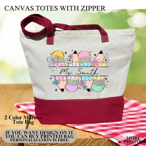 May include: Canvas tote bag with a zipper, featuring a maroon base and handles. The bag has a cream-coloured body with a colourful pencil design and the name "Ms. Smith". The bag is designed for personalisation.