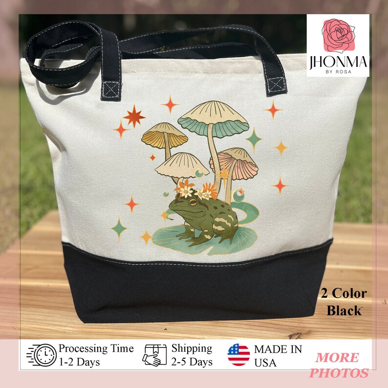 K&ouml;nnte beinhalten: Eine wei&szlig;e Canvas-Tragetasche mit schwarzen Henkeln und einem schwarzen Boden. Die Tasche zeigt ein fantasievolles Design mit einem Frosch, der eine Blumenkrone tr&auml;gt und auf einem Seerosenblatt sitzt, umgeben von Pilzen und Sternen. Der Text "JHONMA BY ROSA" ist in der oberen rechten Ecke der Tasche aufgedruckt.