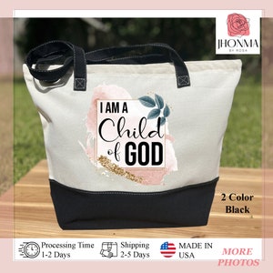 May include: A white canvas tote bag with black handles and a black bottom. The bag has a pink and gold watercolor design with the text "I AM A Child of GOD" in a black square.