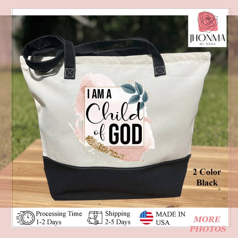 May include: A white canvas tote bag with black handles and a black bottom. The bag has a pink and gold watercolor design with the text "I AM A Child of GOD" in a black square.