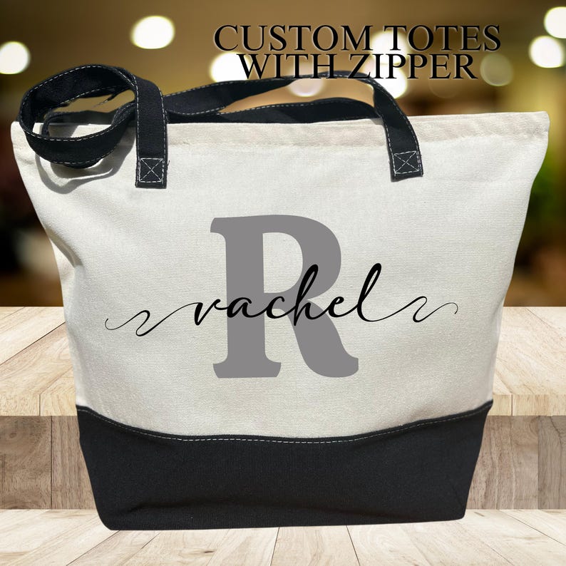 May include: A white canvas tote bag with a black zipper and black handles. The bag has a gray letter "R" and the name "rachel" written in cursive on the front.
