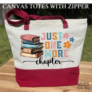 May include: Canvas tote bag with a maroon base and handles, featuring the text "JUST ONE MORE chapter" and a stack of books. The bag has a zipper closure and is designed for carrying books and other items.