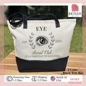 May include: White canvas tote bag with black handles and a black bottom. The bag has a design of an eye with a laurel wreath and the text "EYE Social Club EST. 2024 IT'S A GOOD DAY TO SAVE EYES".