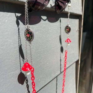 May include: A pair of black sunglasses with a red and white mushroom charm on each side. The sunglasses are hanging from a silver chain with a floral charm.