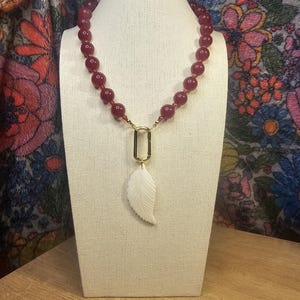 May include: A burgundy beaded necklace with a gold-colored clasp and a white leaf pendant. The necklace is displayed on a mannequin with a floral patterned background. The beads are large and round.