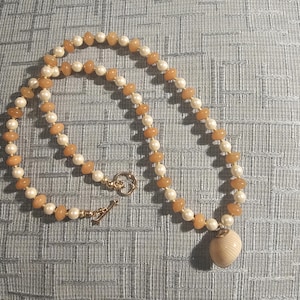 May include: A necklace featuring alternating peach-colored and white beads, with a gold-colored clasp. The necklace has a pendant in the shape of a seashell. The necklace is displayed on a textured, light blue surface.