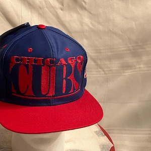 May include: A blue baseball cap with a red brim. The cap features the embroidered text "CHICAGO CUBS" in red. The cap has a structured fit and a curved brim.