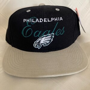 May include: Black and tan baseball cap with the text "PHILADELPHIA Eagles" in white and green script. The cap features the team's logo, an eagle head, on the front. The cap has a flat brim and a snapback closure.