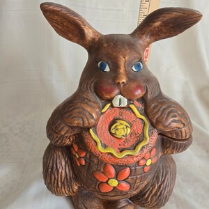 May include: A brown ceramic bunny cookie jar with blue eyes and a red and yellow flower design. The bunny is holding a red and yellow donut in its paws.