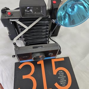 May include: A black Polaroid Land Camera Model 315 with a blue flash attachment. The camera has a red button on the top and a black instruction booklet with the text "How to Use Your Polaroid Land Camera Model 315".