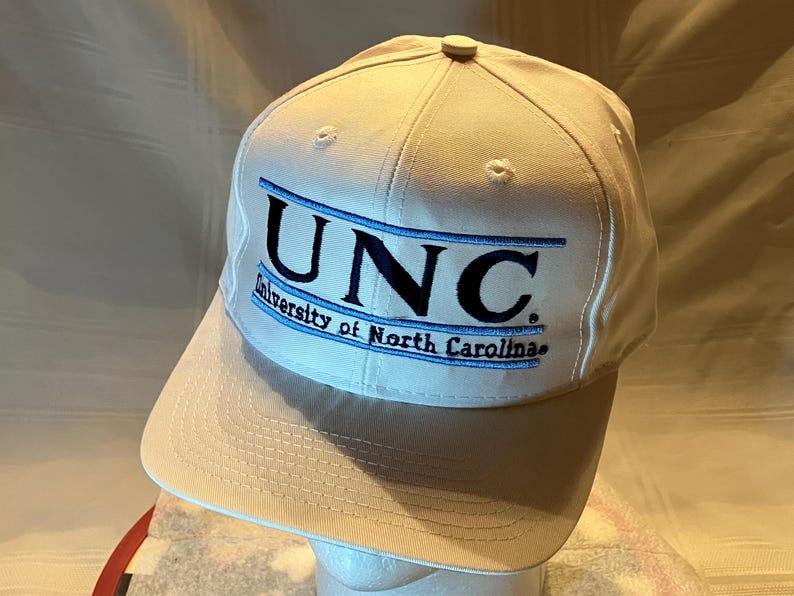 May include: A white baseball cap with the letters "UNC" embroidered in black and blue. The words "University of North Carolina" are embroidered below the letters. The cap has a curved brim and a button on top.