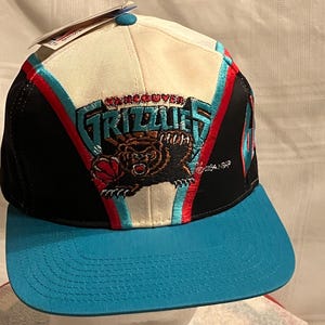 May include: A vintage Vancouver Grizzlies hat with a teal brim and a white crown. The hat features the team's logo, a bear holding a basketball, embroidered in teal, red, and brown. The hat has a black side panel and a tag attached.