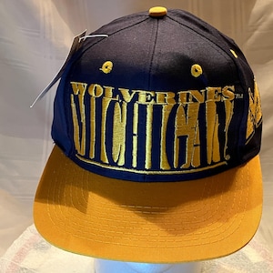 May include: A navy blue and maize yellow baseball cap. The cap features the word "Wolverines" arched above "Michigan" in gold embroidery. The brim is maize yellow, and the top of the cap is navy blue.