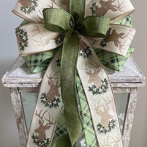 May include: A decorative Christmas bow featuring a deer design with gold glitter accents and holly. The bow is made of cream and green plaid ribbon, with a large green bow in the center. The bow is placed on a white lantern.