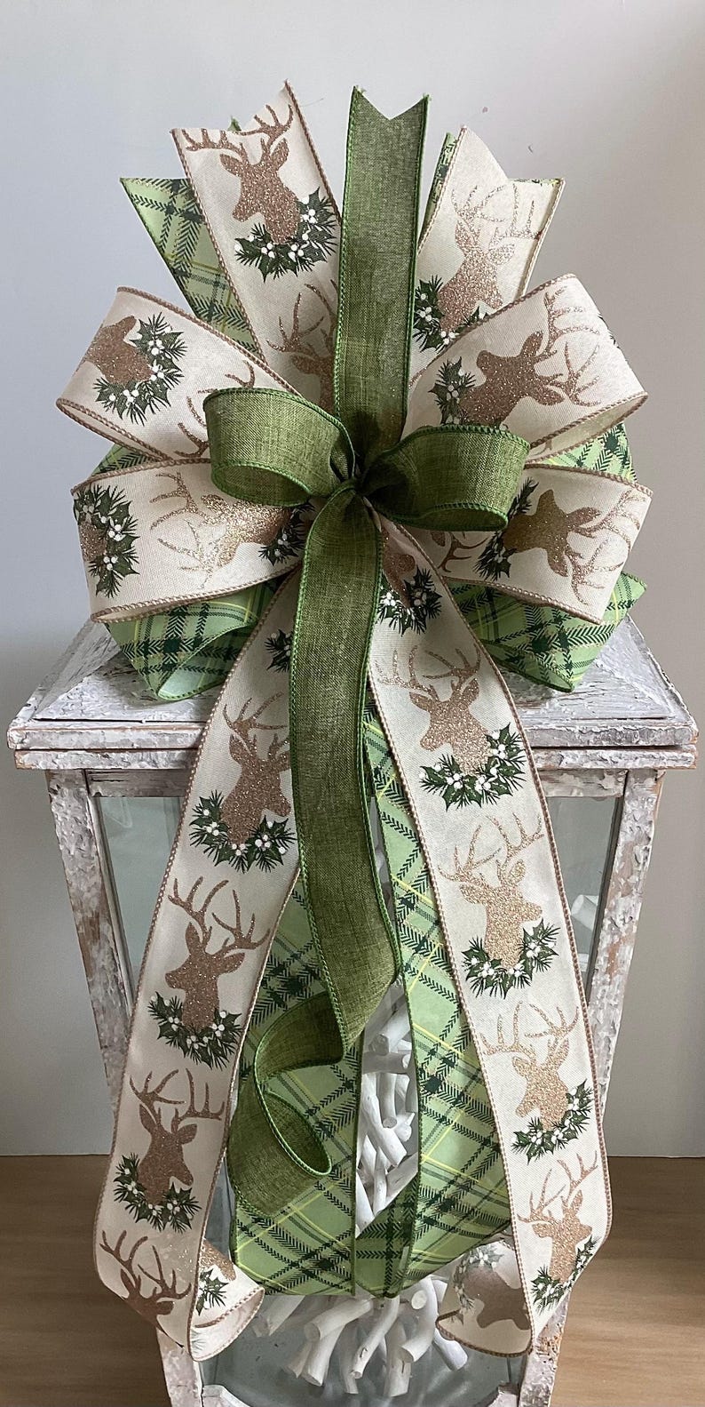 May include: A decorative Christmas bow featuring a deer design with gold glitter accents and holly. The bow is made of cream and green plaid ribbon, with a large green bow in the center. The bow is placed on a white lantern.