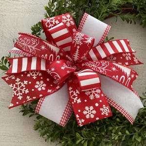 Pode incluir: Um laço de Natal festivo com fitas vermelhas e brancas adornadas com motivos de flocos de neve, listras e árvores. O laço é colocado sobre um fundo de folhagem verde, criando um acento decorativo sazonal. O design inclui uma mistura de texturas e padrões.