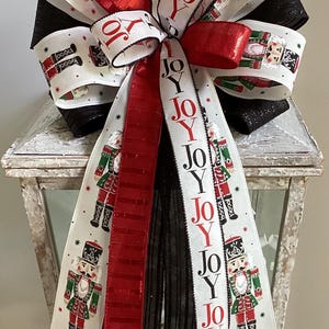 May include: A decorative Christmas bow featuring a festive design. The bow has multiple layers of white, red, and black ribbons. The white ribbons display a nutcracker soldier and the word "JOY". The bow is placed on a white lantern.