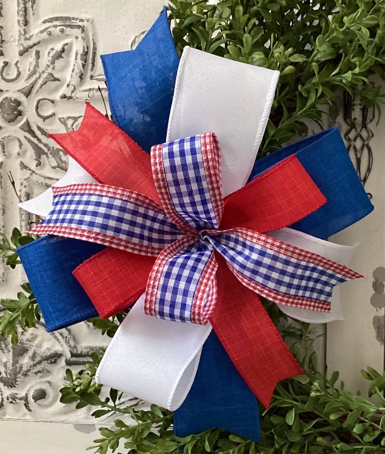 May include: A red, white, and blue bow with a gingham pattern. The bow is made of ribbon and is tied in a knot. The bow is on a green background.
