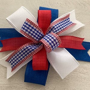May include: A red, white, and blue bow made with gingham ribbon and solid fabric ribbon. The bow is tied in a knot and has a layered look.