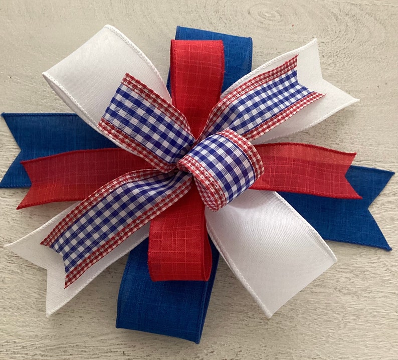 May include: A red, white, and blue bow made with gingham ribbon and solid fabric ribbon. The bow is tied in a knot and has a layered look.