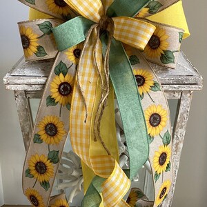 Könnte beinhalten: Eine weiße Laterne mit einer Sonnenblumen-Schleife aus Sackleinen, gelbem Gingham und grünem Band. Die Schleife ist mit Schnur gebunden und die Laterne hat ein Glasfenster.