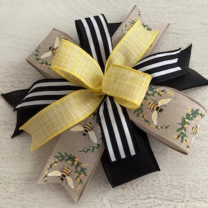 May include: A decorative bow made with black and white striped ribbon, yellow gingham ribbon, and beige ribbon with a bee and floral pattern.