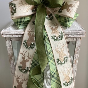 May include: A decorative Christmas bow featuring a layered design with green plaid and cream-colored ribbons. The cream ribbons display a gold glitter deer and holly design. A large green bow is at the center.