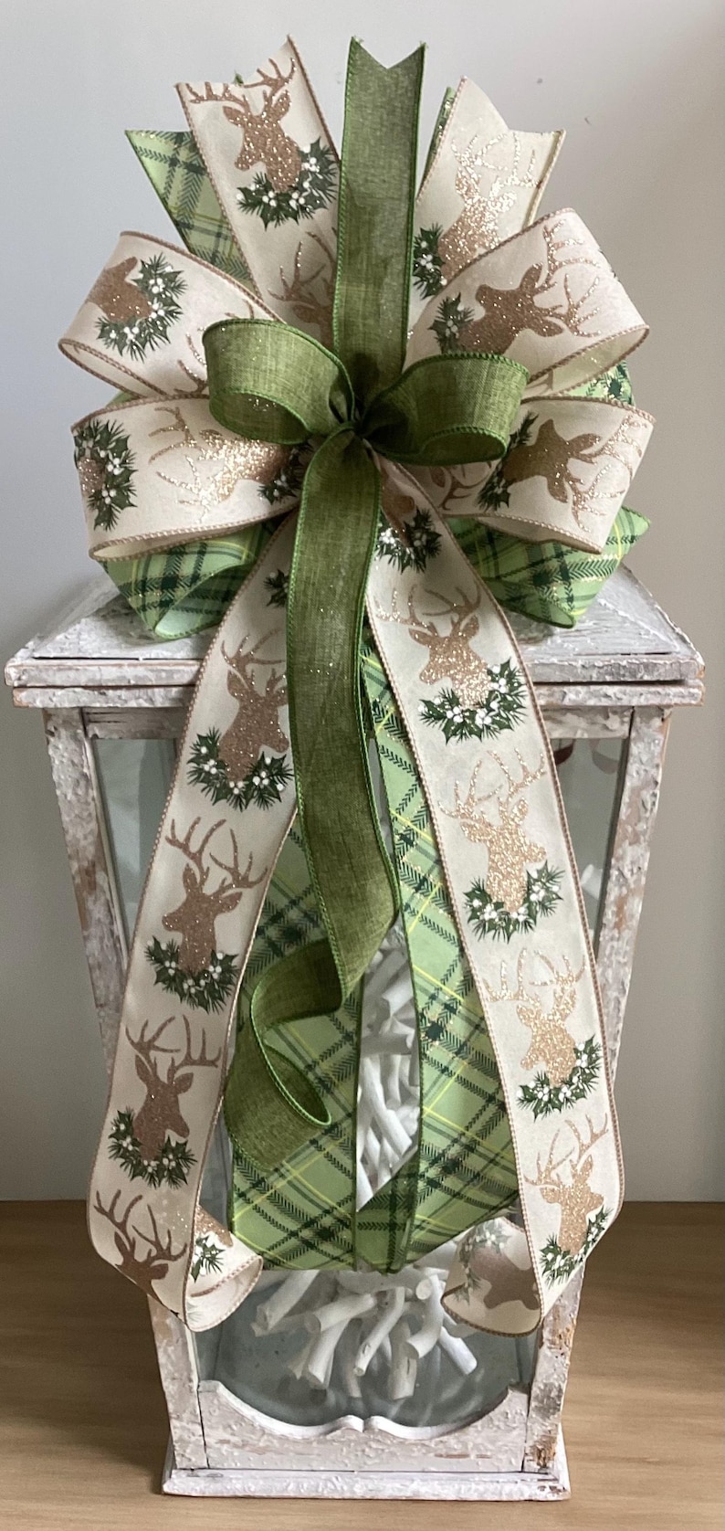 May include: A decorative Christmas bow featuring a layered design with green plaid and cream-colored ribbons. The cream ribbons display a gold glitter deer and holly design. A large green bow is at the center.