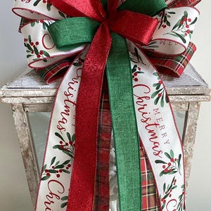 May include: A Christmas bow in red, green, and white. The bow has a large red bow, green and red plaid ribbons, and white ribbons printed with "Merry Christmas" and holly berry sprigs. The bow is placed on a white lantern.