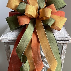 Könnte beinhalten: Dekorative Schleife mit Herbstfarben: Orange, Grün und Gold. Die Schleife besteht aus strukturiertem Band und ist in einem geschichteten Design angeordnet. Die Schleife befindet sich auf einer weißen Laterne.
