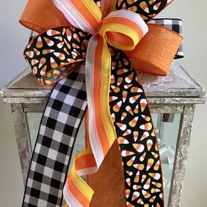 May include: A decorative Halloween bow featuring multiple ribbons in orange, black, white, and yellow. The bow includes ribbons with candy corn patterns, a black and white buffalo check, and solid orange. The bow is attached to a white lantern.