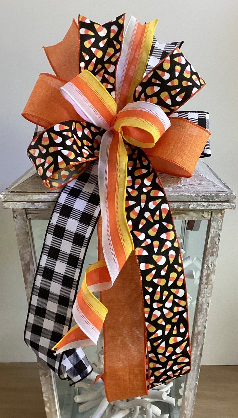 May include: A decorative Halloween bow featuring multiple ribbons in orange, black, white, and yellow. The bow includes ribbons with candy corn patterns, a black and white buffalo check, and solid orange. The bow is attached to a white lantern.