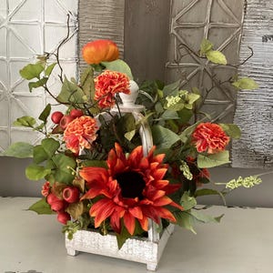 May include: A white wooden basket overflowing with vibrant orange and red artificial flowers, including a large sunflower and smaller blooms. Green leaves and berries add to the arrangement. The basket has a handle and sits on a white surface.