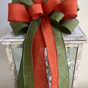 May include: A decorative bow featuring layers of orange and green fabric. The bow is placed on top of a white, distressed lantern. The bow's design includes multiple loops and long, flowing ribbons, creating a festive and elegant look.
