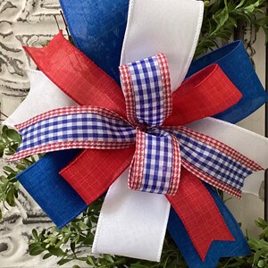 May include: A red, white, and blue bow made with gingham and burlap ribbon. The bow is tied around a wreath of green boxwood.