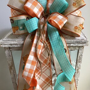 May include: A fall-themed bow made with three different ribbons. The ribbons are a light blue plaid, a white and orange plaid, and a beige ribbon with a vintage truck and pumpkins design. The bow is tied with twine and is displayed on a white lantern.