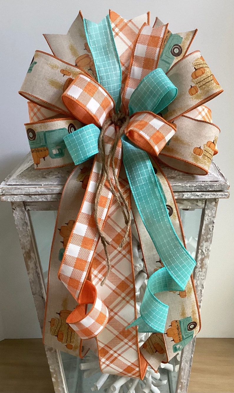 May include: A fall-themed bow made with three different ribbons. The ribbons are a light blue plaid, a white and orange plaid, and a beige ribbon with a vintage truck and pumpkins design. The bow is tied with twine and is displayed on a white lantern.