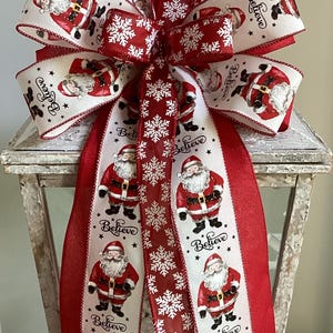 May include: A festive Christmas bow with a red and white design. The bow has a large, multi-loop shape with ribbons printed with Santa Claus illustrations and the word "Believe". Snowflake patterns add to the holiday theme.