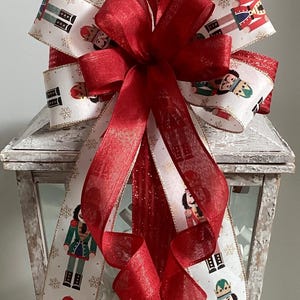 May include: A red and white Christmas bow with a nutcracker pattern. The bow is tied with a red ribbon and is decorated with gold snowflakes. The bow is sitting on top of a white lantern.